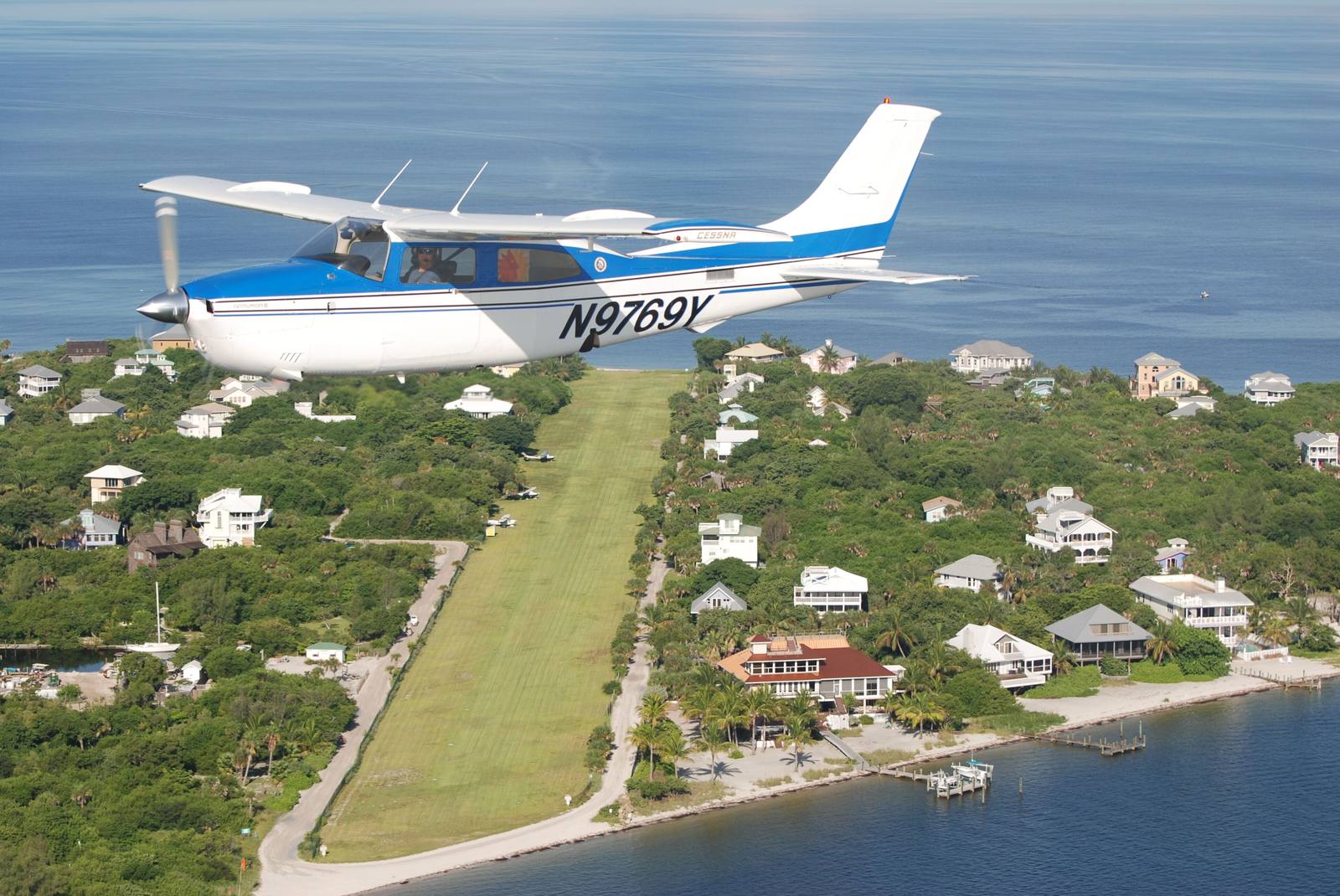 A small plane flying over the barrier island, with the grass airstrip and houses below